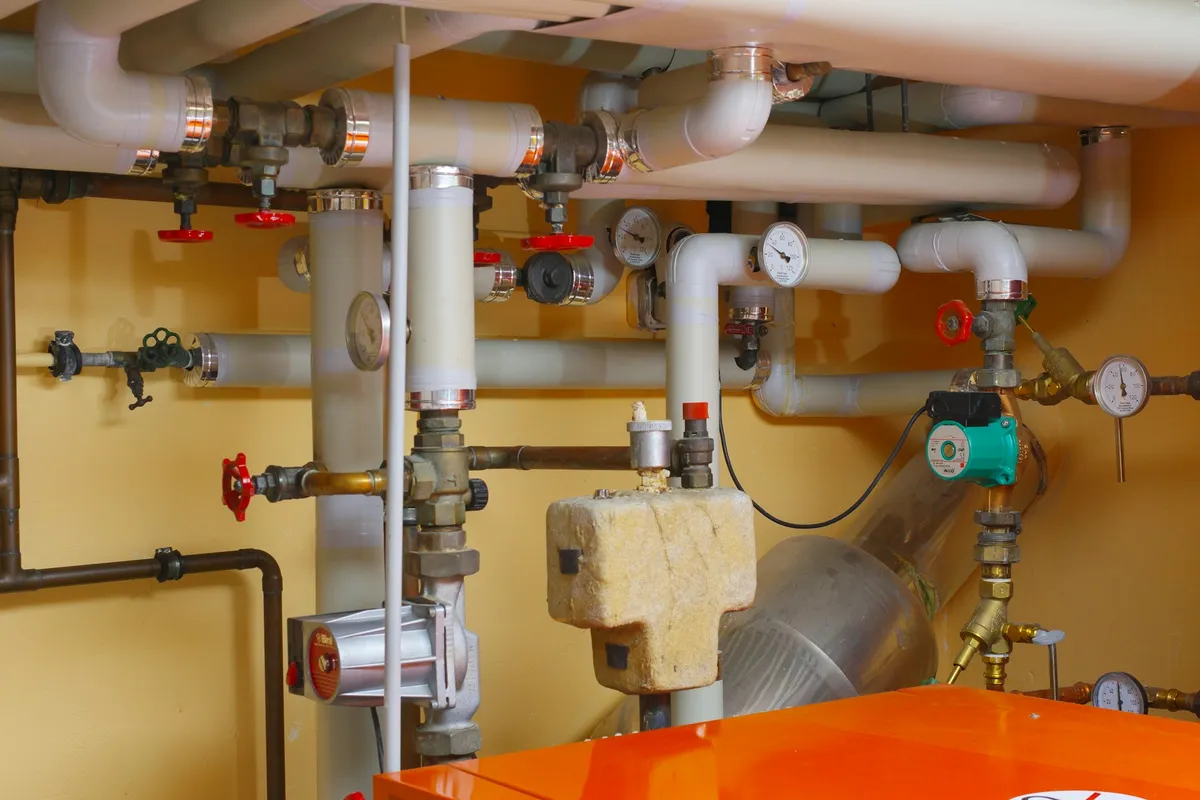 Boiler room wall with insulated pipes, valves and pressure gauges above an orange boiler