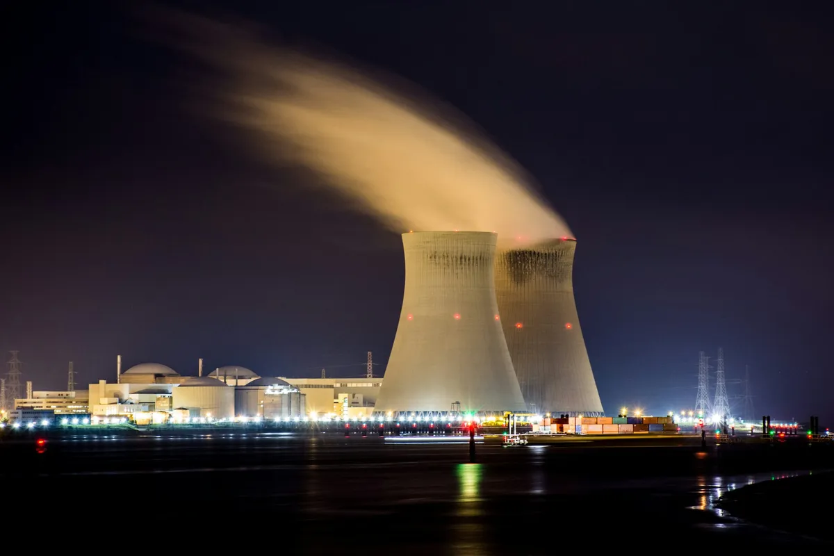 Two large cooling towers releasing a long plume of steam at night beside a lit power station and a dark river