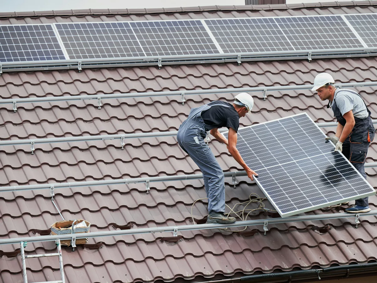 Two installers in hard hats lifting a solar panel into place on a tiled house roof
