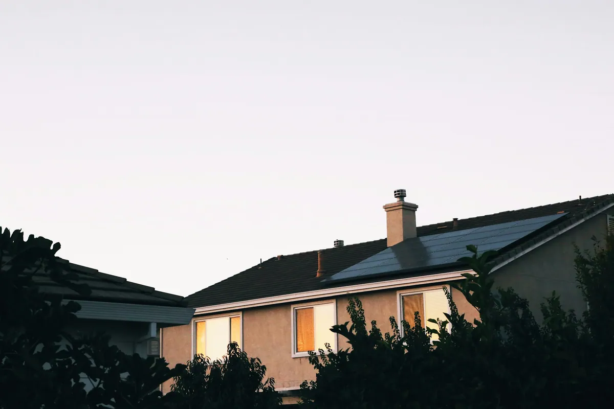 House at dusk with warm lit windows and solar panels on the roof under a pale evening sky