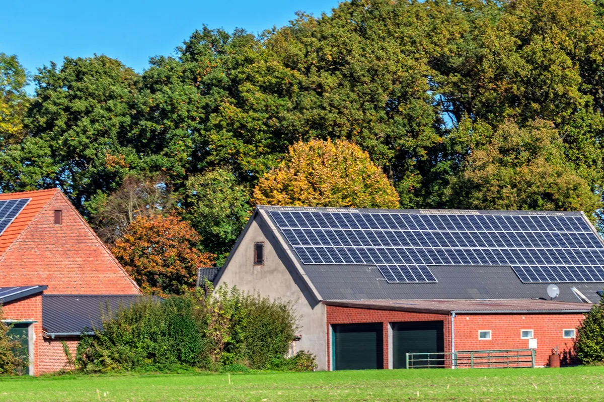 Barn roof covered in solar panels beside red-brick farm buildings and autumn trees