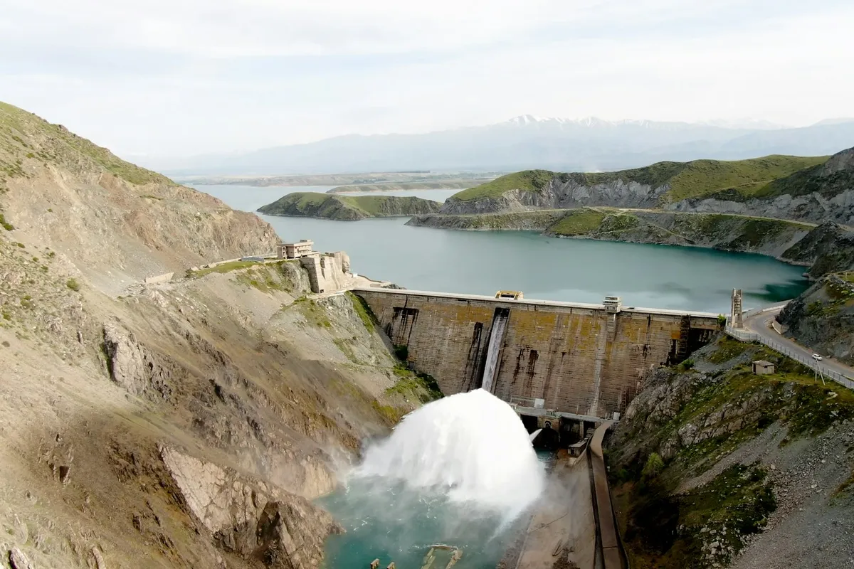 Concrete dam with white water rushing from its spillway into a river below a reservoir