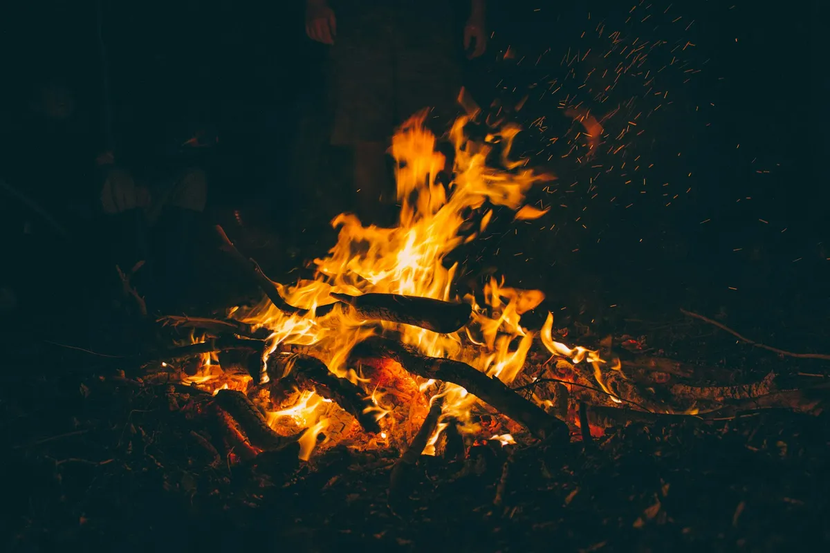 Open campfire at night with bright orange flames, glowing embers and sparks rising above burning logs