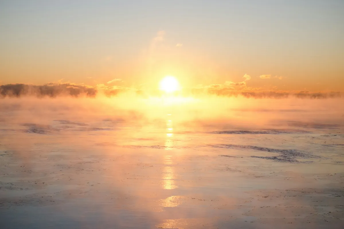 Low sun glowing orange over a misty, ice-flecked water surface with a reflected column of light