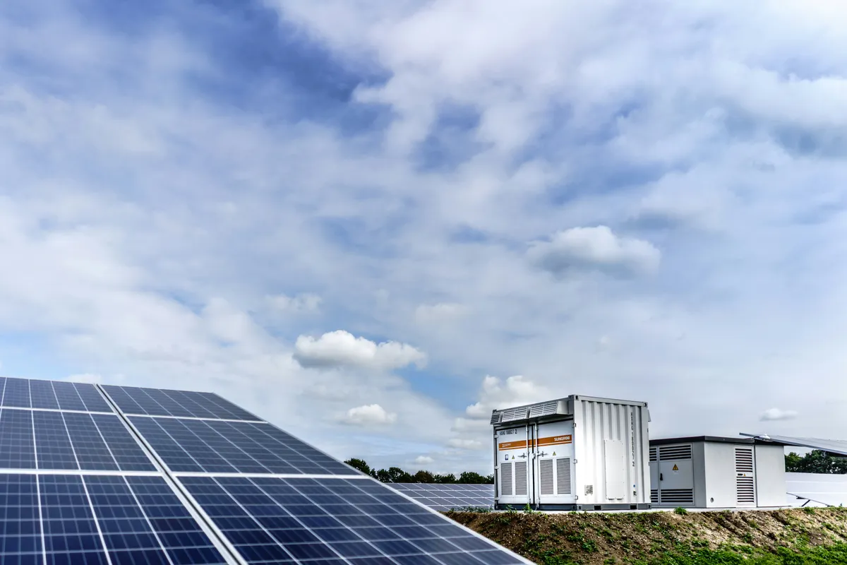 Ground-mounted solar array beside a white battery energy storage container under a cloudy sky